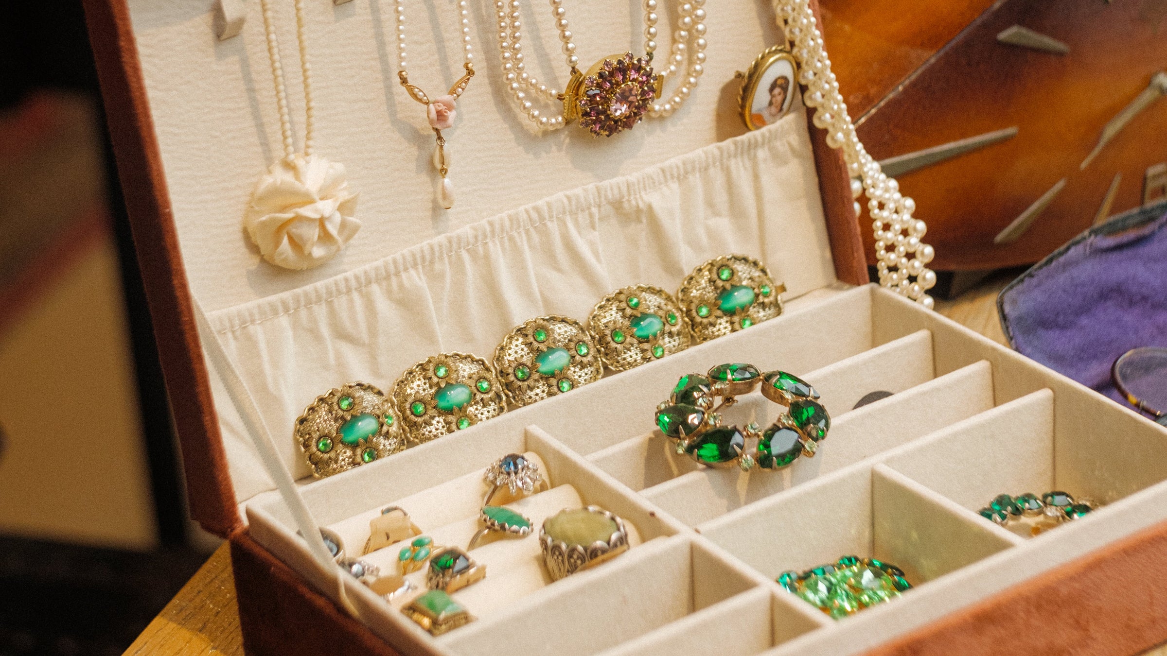 An open brown jewelry box with cream interior compartments, holding pearl necklaces, beaded strands, and multiple green-and-gold brooches and rings, set on a wooden table with a softly blurred yellow hat in the background.